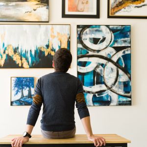 Hispanic male visitor looking reflective while sitting on a bench and admiring the various paintings on the wall of an art gallery